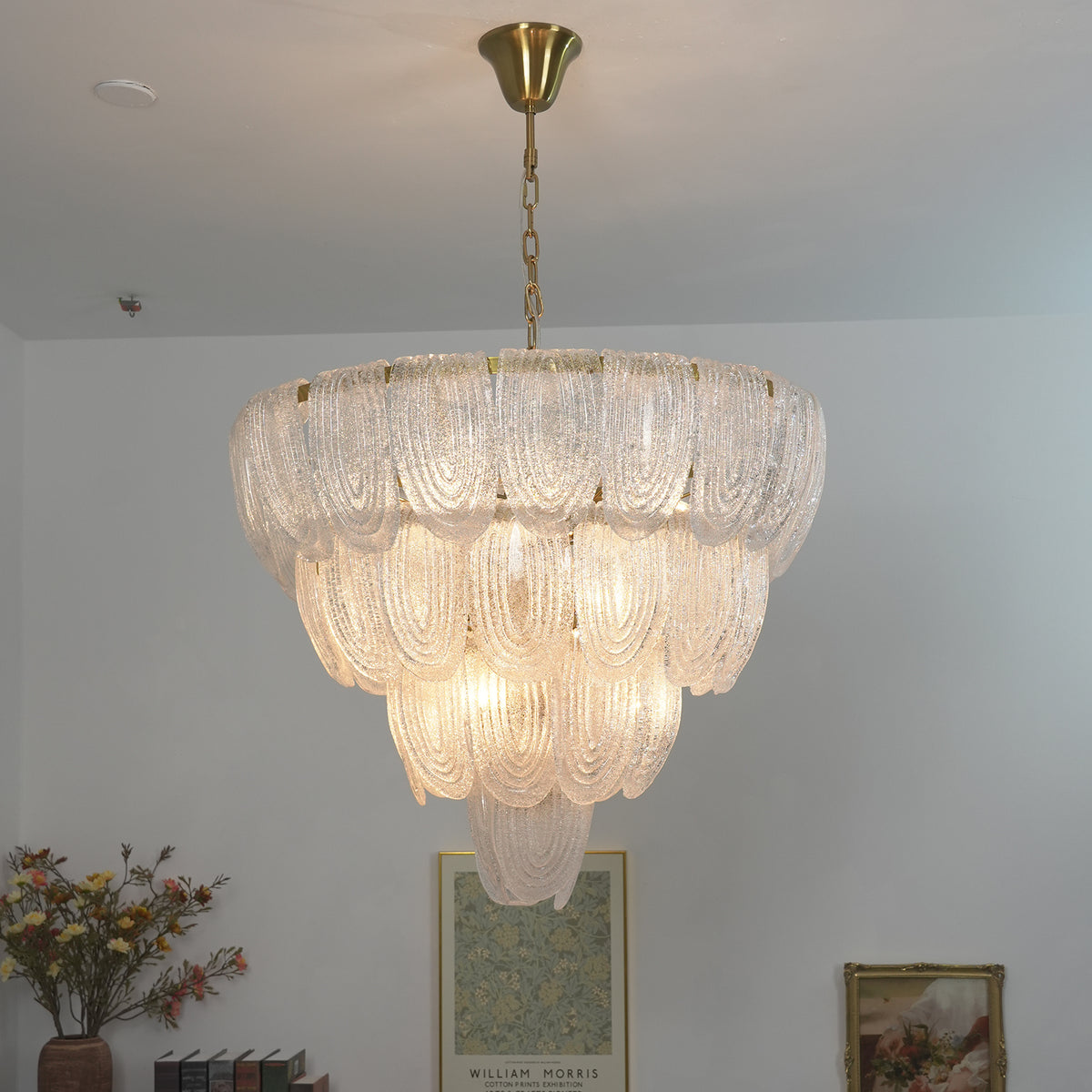 Lit layered glass petal chandelier casting a soft warm glow against a light wall background.