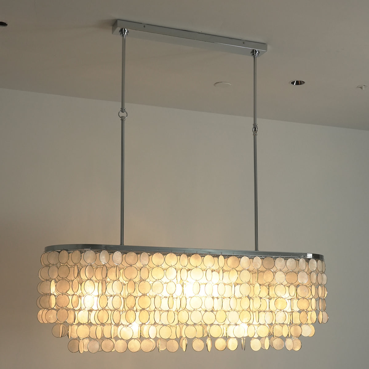 Lit rectangular capiz shell chandelier casting a soft warm glow against a neutral wall background.