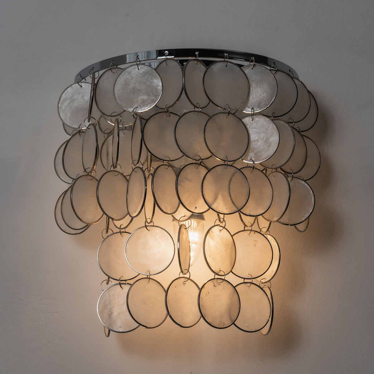 Front view of a lit round tiered capiz shell wall sconce, highlighting its cascading shell layers.