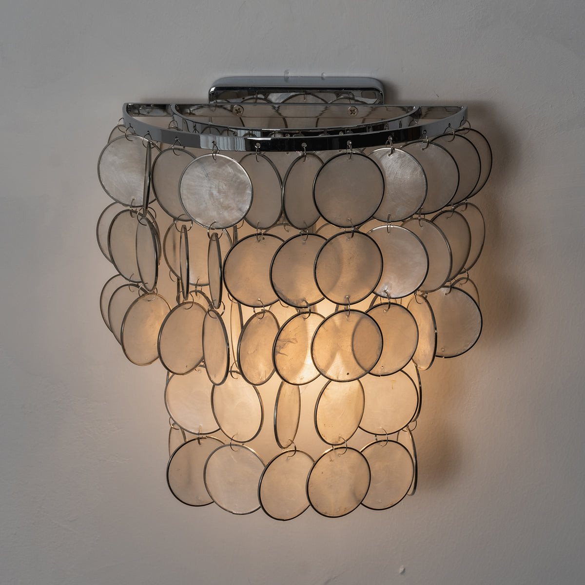 Close-up of a lit round tiered capiz shell wall sconce, showing warm light filtering through natural shells.