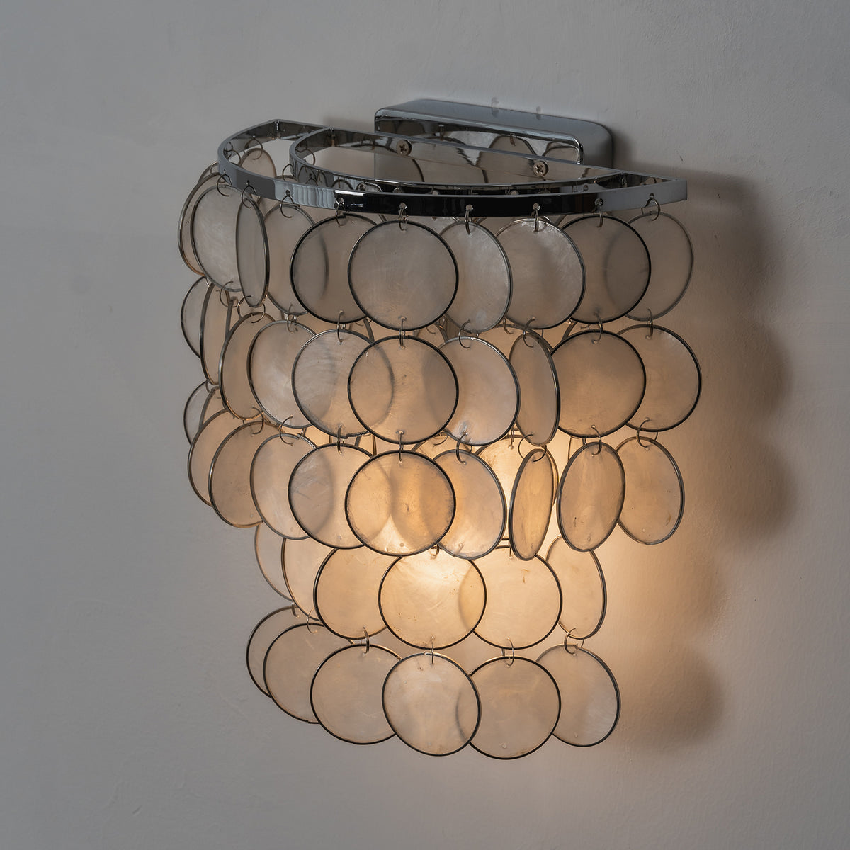 Lit round tiered capiz shell wall sconce with soft warm glow against a neutral wall background.