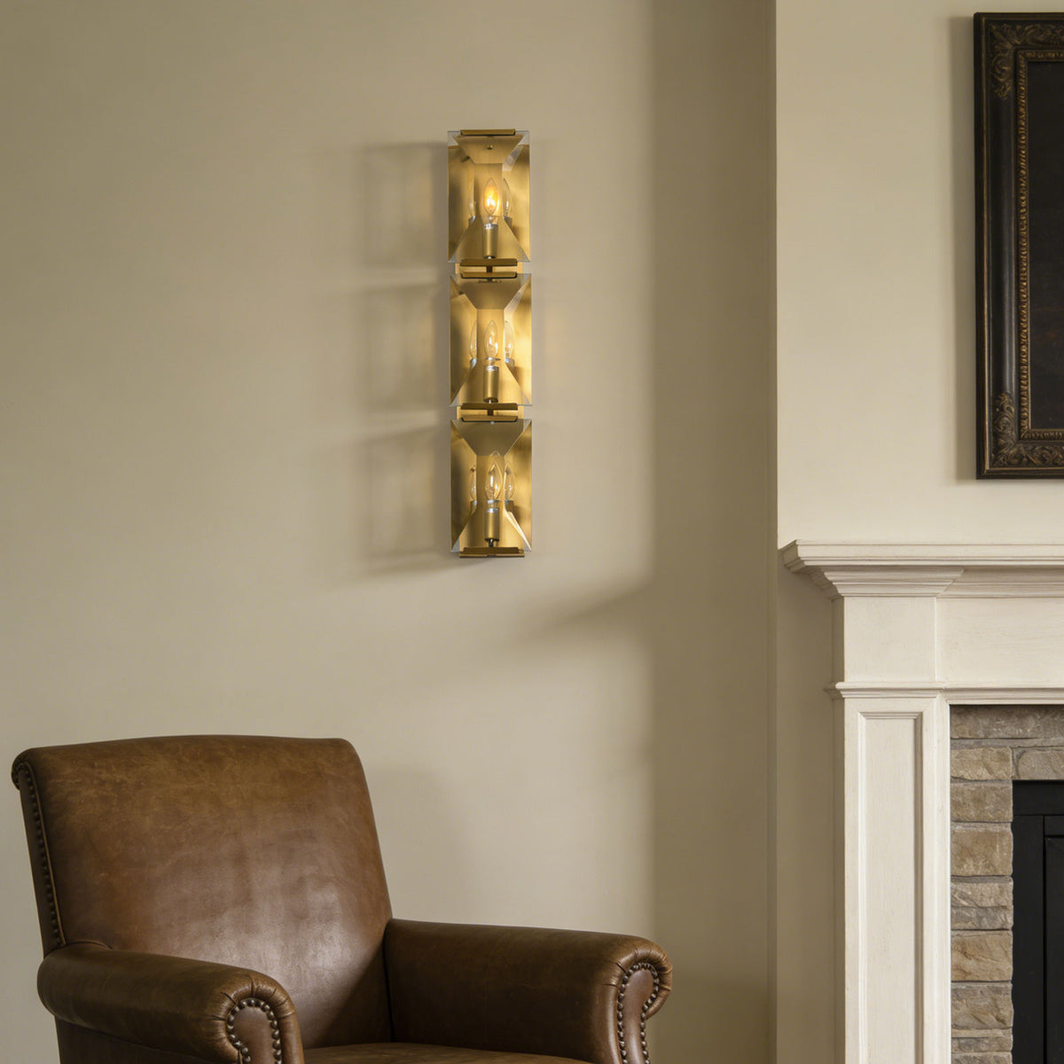 Three-headed crystal wall lamp beside a brown leather armchair and fireplace in a cozy living room.