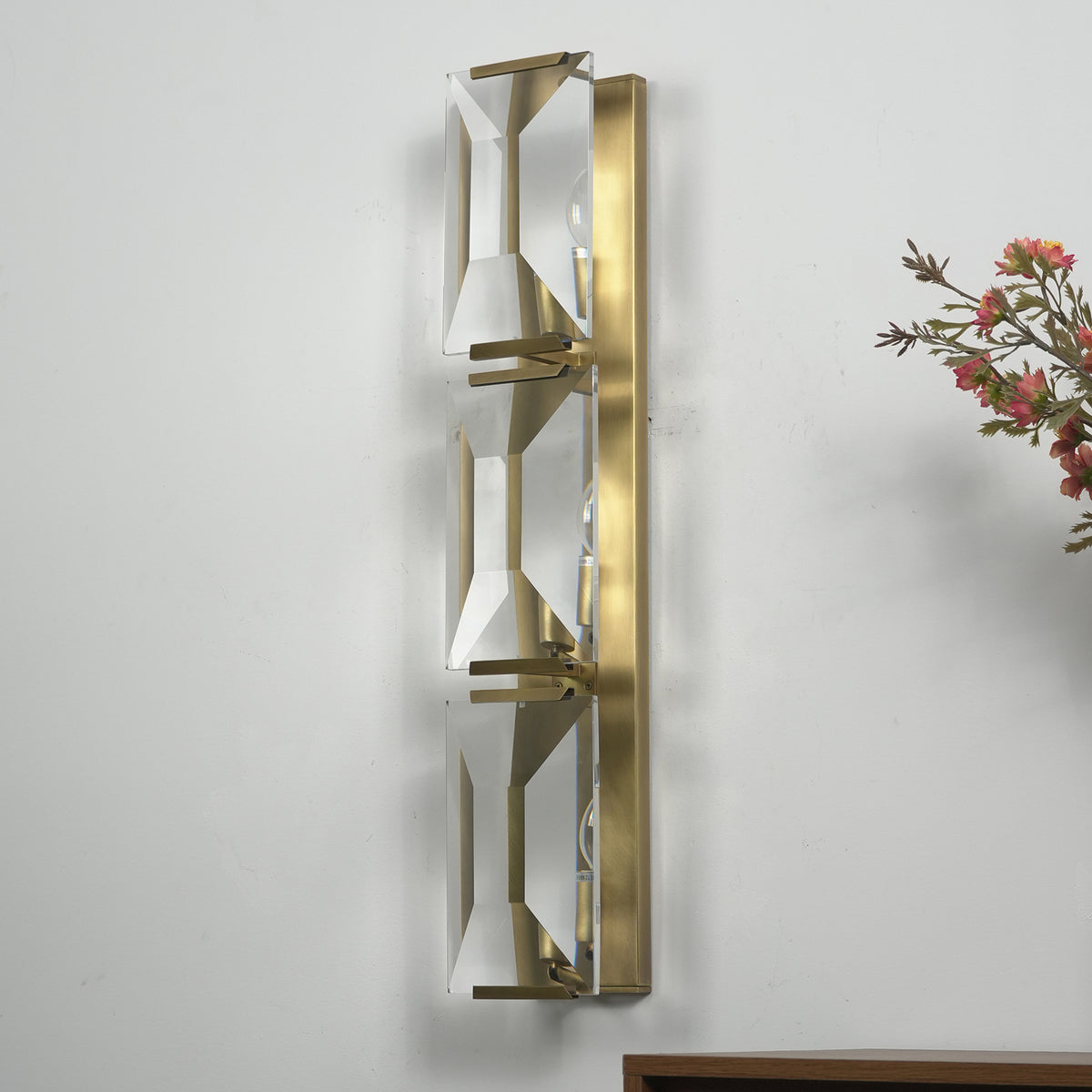 Three-headed crystal wall lamp mounted on a light wall, casting warm light in a modern room.