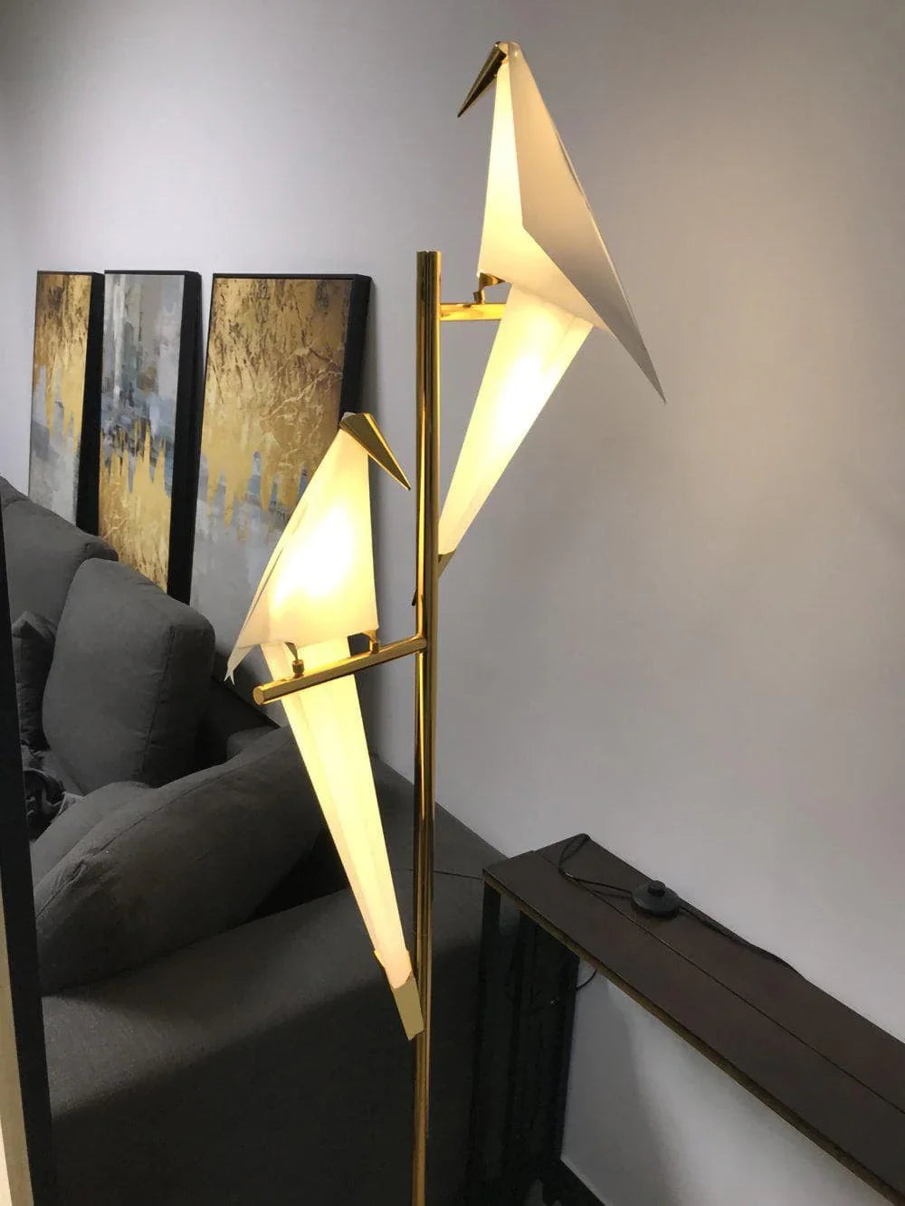 Modern origami bird floor lamp with gold finish, glowing warmly in living room setting