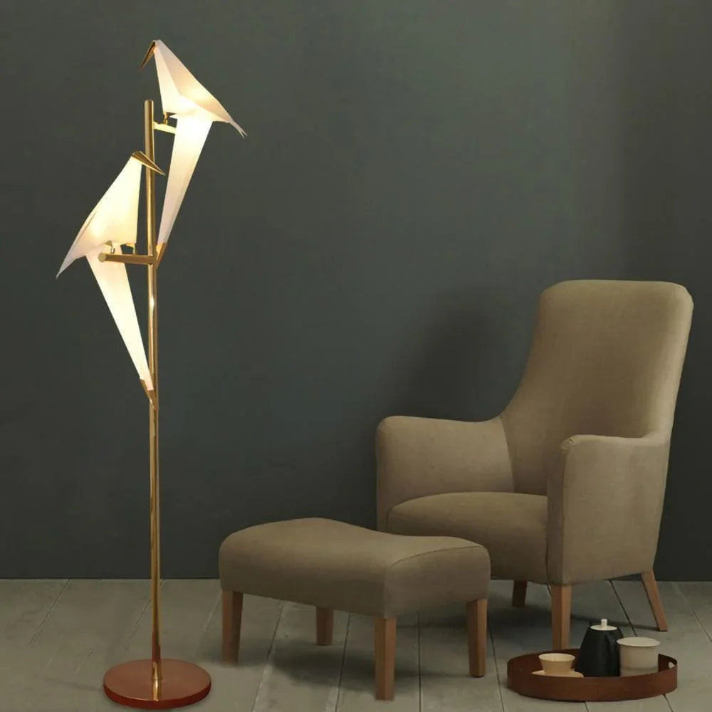 Modern bird-shaped floor lamp with gold stand, illuminating a cozy living room corner with armchair and ottoman