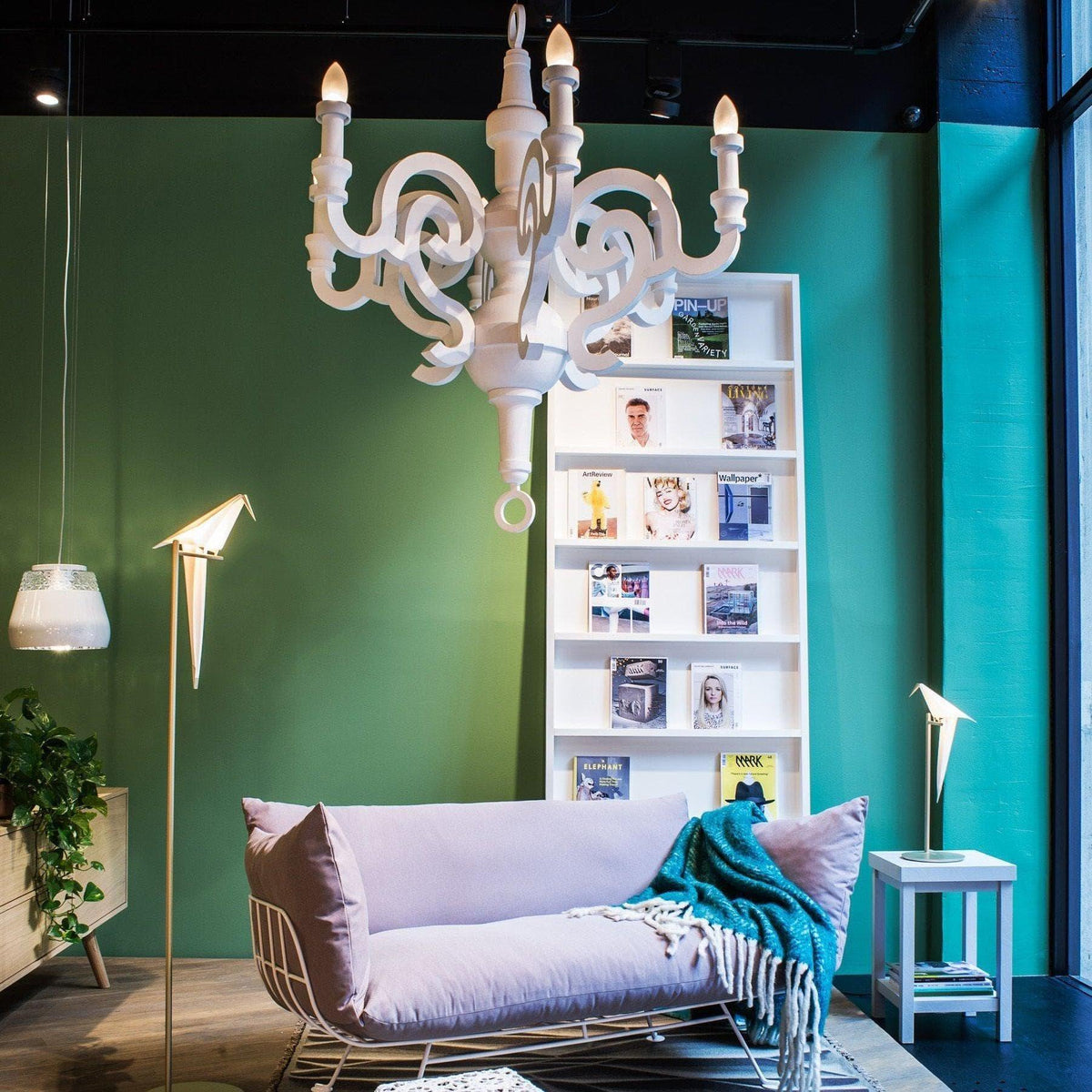 White ornate chandelier over modern lavender sofa in stylish living room with green walls and magazine shelf
